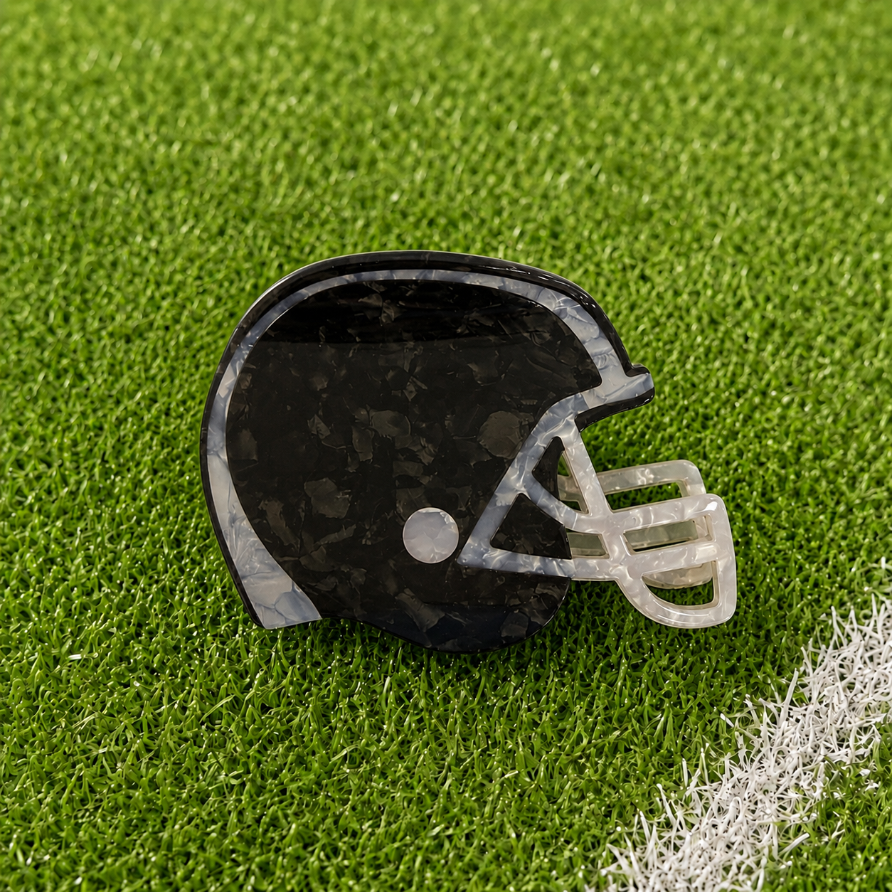 A football helmet-shaped hair claw with a black and gray marbled pattern and a translucent white faceguard, resting on green artificial turf.