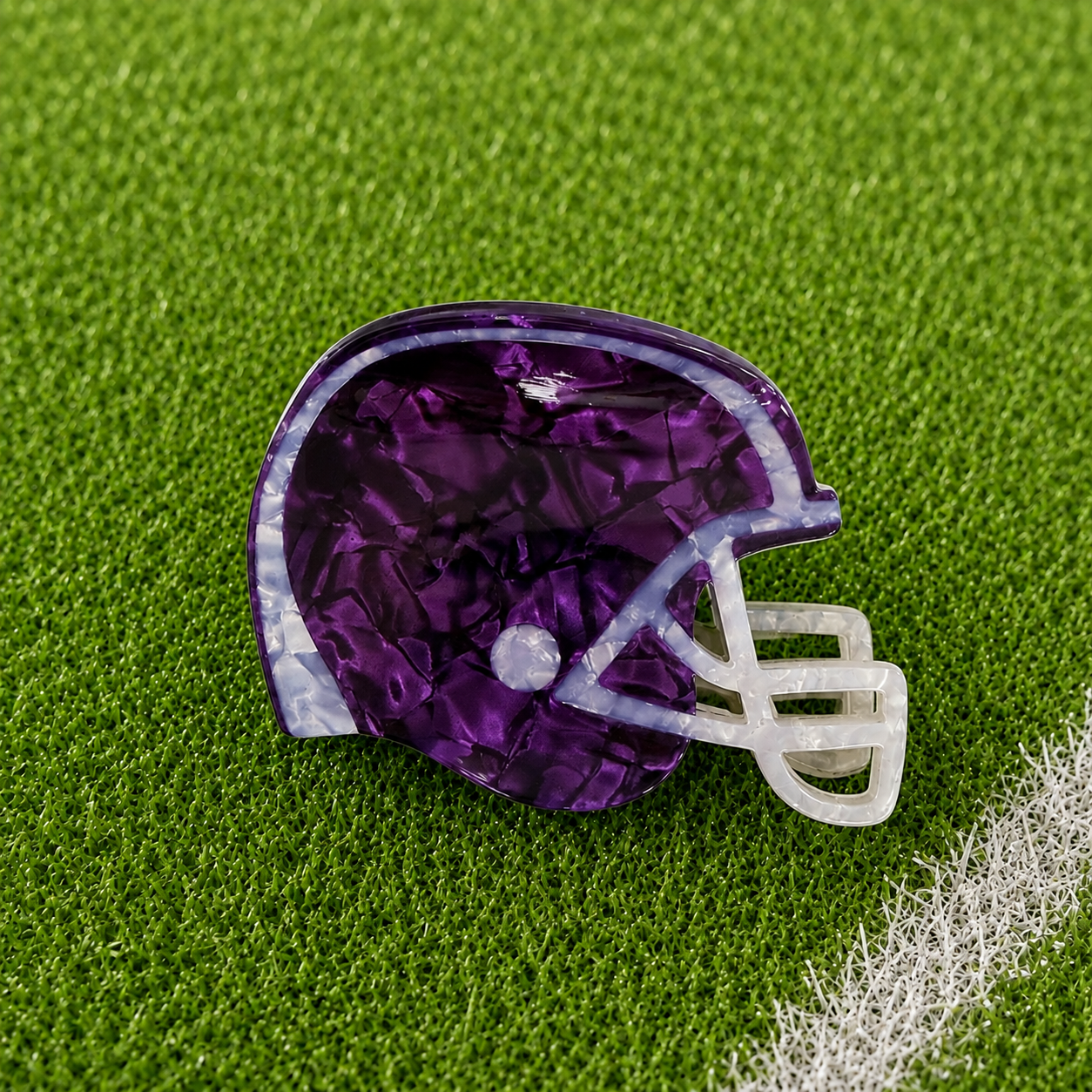 A purple football helmet-shaped hair claw with a white faceted face guard, resting on green artificial turf.