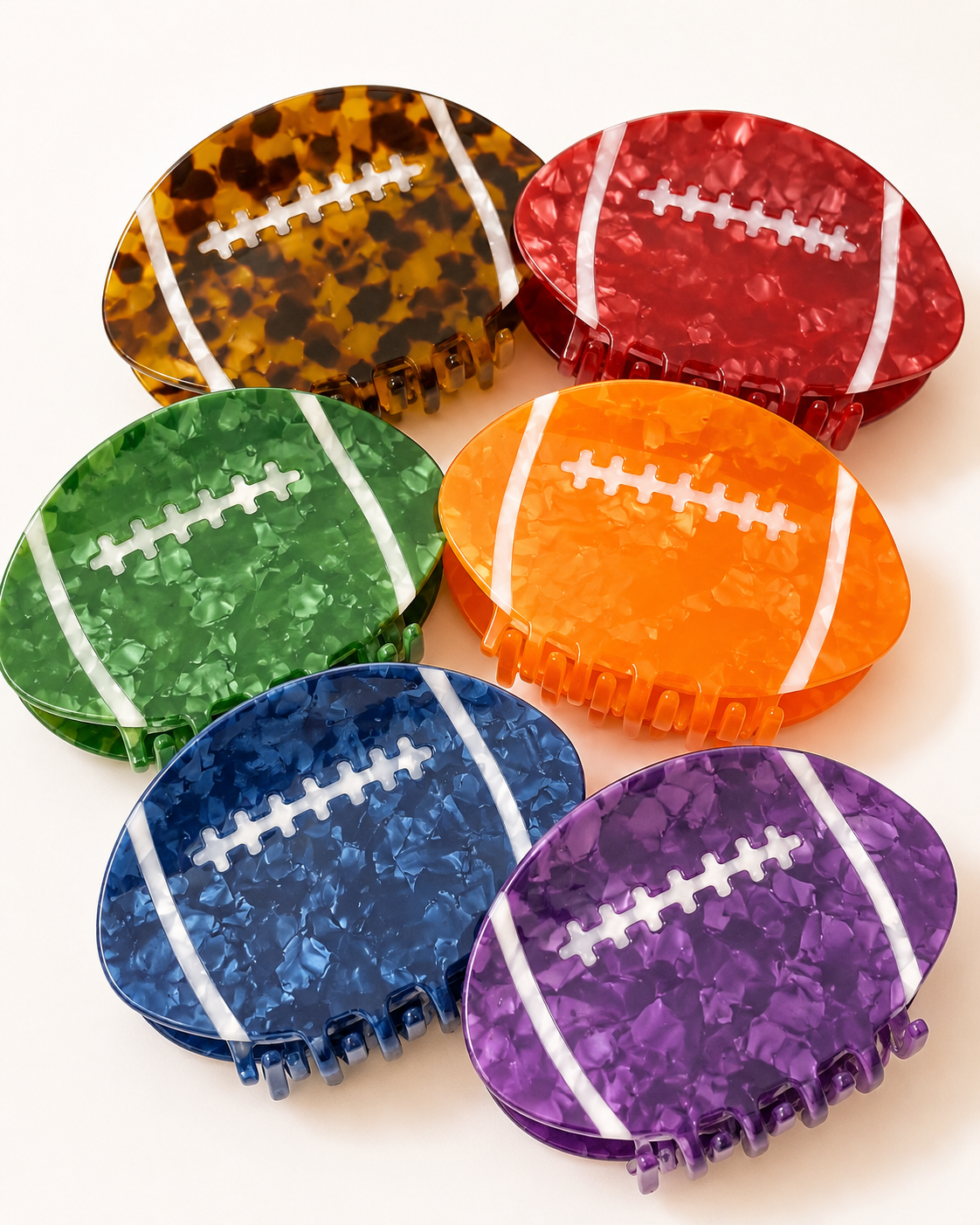 Six football-shaped hair claw clips in assorted colors, including tortoiseshell, red, green, orange, blue, and purple, arranged on a light cream background.
