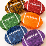 Six football-shaped hair claw clips in assorted colors, including tortoiseshell, red, green, orange, blue, and purple, arranged on a light cream background.