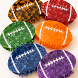 Six football-shaped hair claw clips in assorted colors, including tortoiseshell, red, green, orange, blue, and purple, arranged on a light cream background.