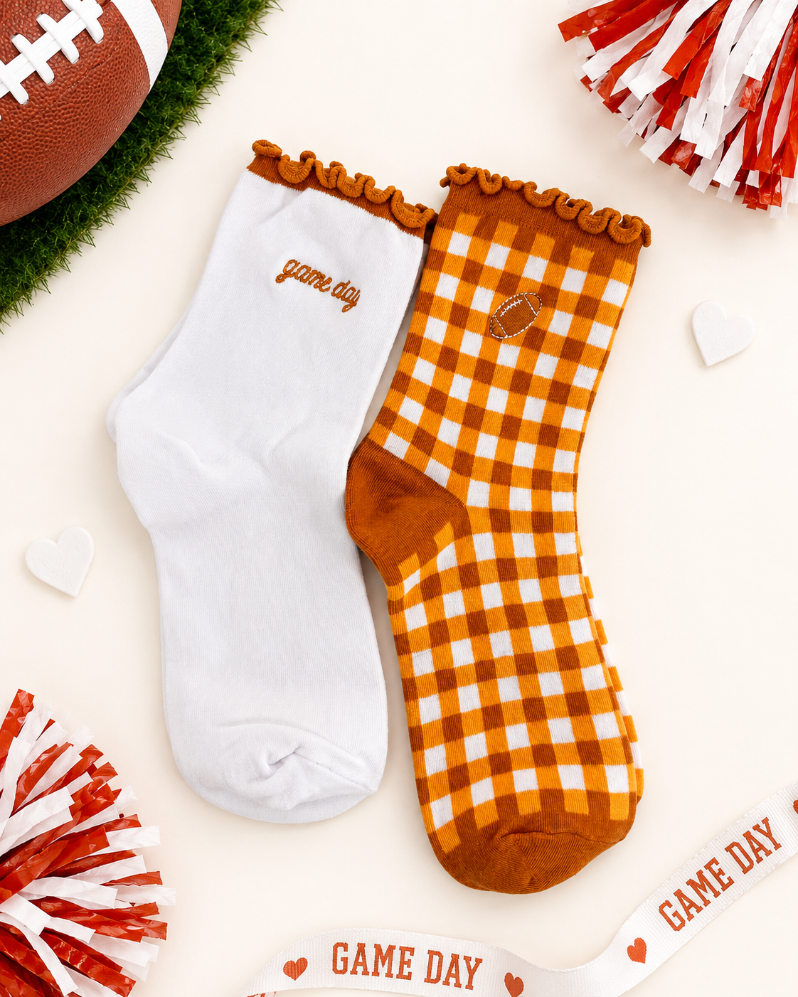 Two decorative ankle socks displayed on a light cream background with football-themed props. One sock is solid white with 'game day' embroidered in brown near the cuff.