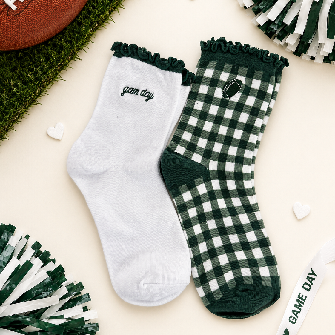 A pair of decorative crew socks laid flat on a cream background with football-themed props. One sock is white with dark green ruffle trim and the word 'game day' in script; the other is dark green and white gingham with a small football patch and 'game day' text.