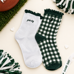 A pair of decorative crew socks laid flat on a cream background with football-themed props. One sock is white with dark green ruffle trim and the word 'game day' in script; the other is dark green and white gingham with a small football patch and 'game day' text.