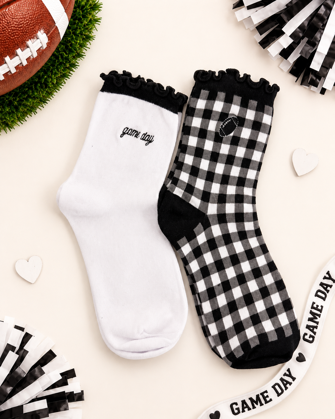 Two socks displayed flat on a light beige background with football-themed props. One sock is solid white with black ruffle trim and 'game day' text, and the other is black-and-white gingham with black ruffle trim, black toe/heel areas, and a small football patch.