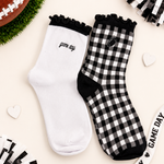 Two socks displayed flat on a light beige background with football-themed props. One sock is solid white with black ruffle trim and 'game day' text, and the other is black-and-white gingham with black ruffle trim, black toe/heel areas, and a small football patch.