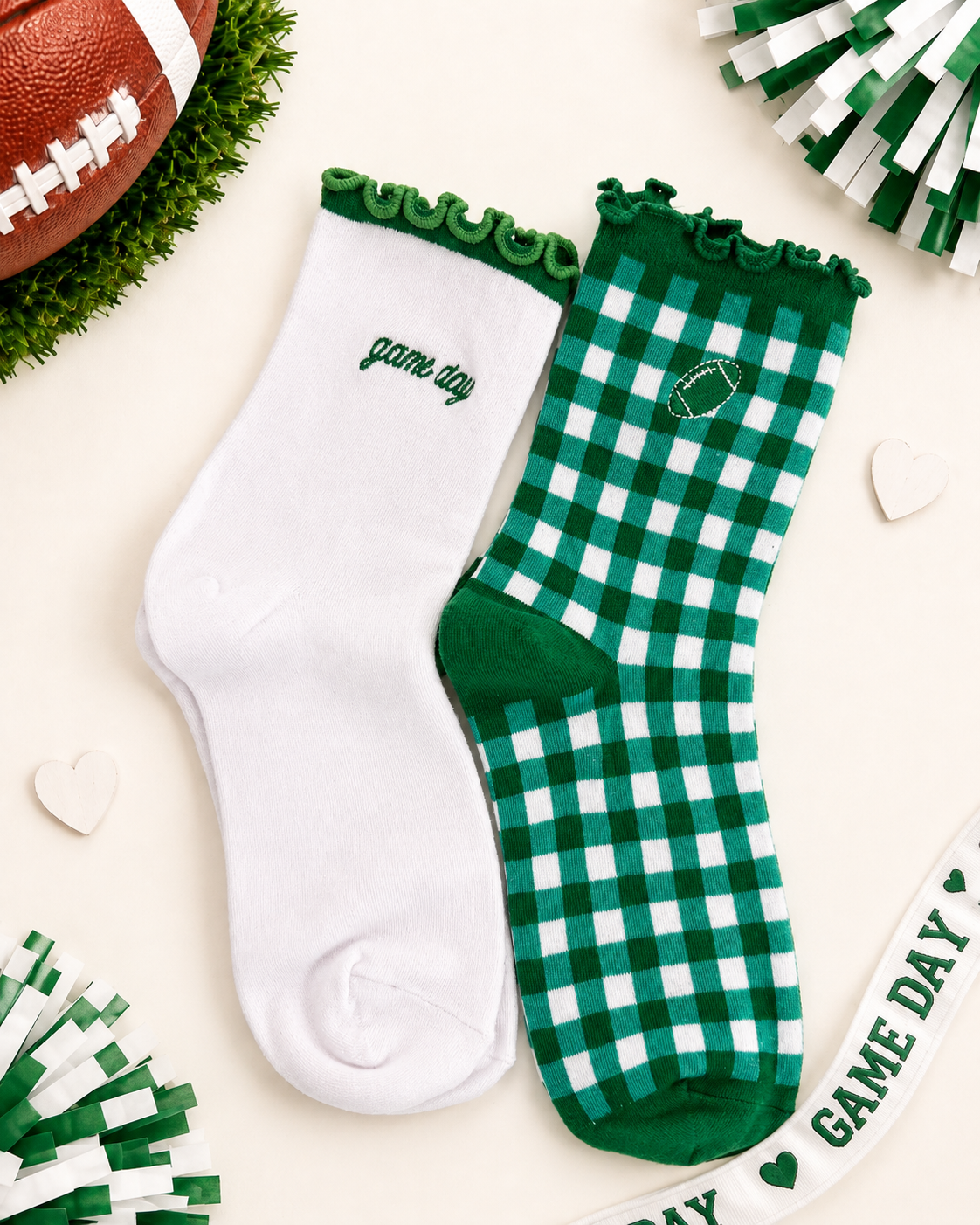 Two socks laid flat on a light cream background with football-themed props. One sock is solid white with green trim and the word 'game day' embroidered near the cuff.