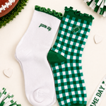 Two socks laid flat on a light cream background with football-themed props. One sock is solid white with green trim and the word 'game day' embroidered near the cuff.