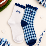 Two pairs of women's crew socks styled in a flat-lay arrangement. One pair is solid white with a navy blue ruffled cuff and 'game day' text on the side.