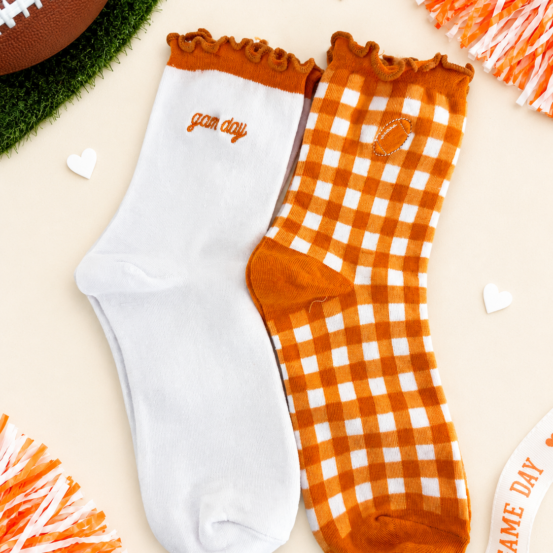 Two crew socks displayed flat on a light beige background with football-themed props. One sock is solid white with orange trim and the word 'game day' embroidered on the side.