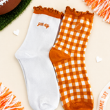 Two crew socks displayed flat on a light beige background with football-themed props. One sock is solid white with orange trim and the word 'game day' embroidered on the side.