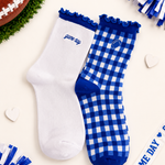 Two socks displayed on a cream background with football-themed props. One sock is solid white with royal blue trim and the embroidered text 'game day'.