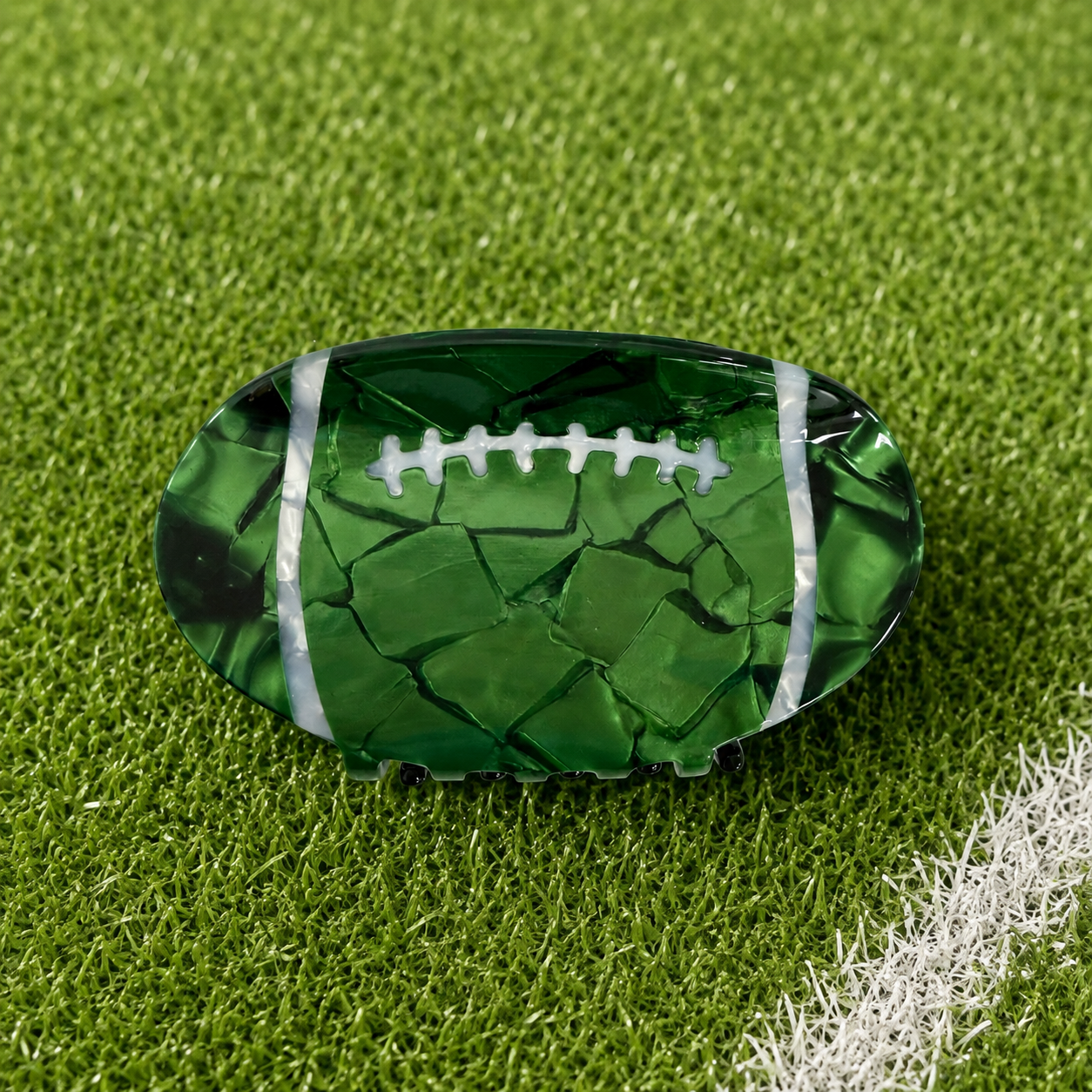 A green football-shaped hair claw clip with a cracked-mercury glass texture and white laces, displayed on a green turf field background.