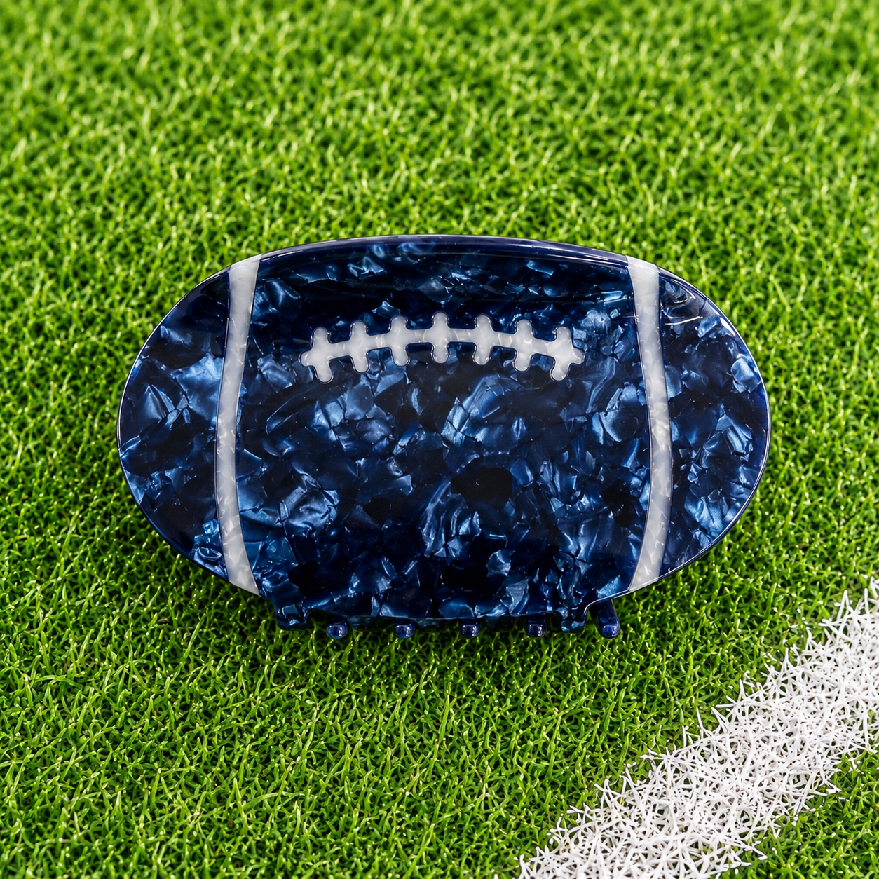 A football-shaped hair claw clip with a navy blue marbled pattern and white laces, displayed on a green grass background.