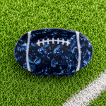 A football-shaped hair claw clip with a navy blue marbled pattern and white laces, displayed on a green grass background.