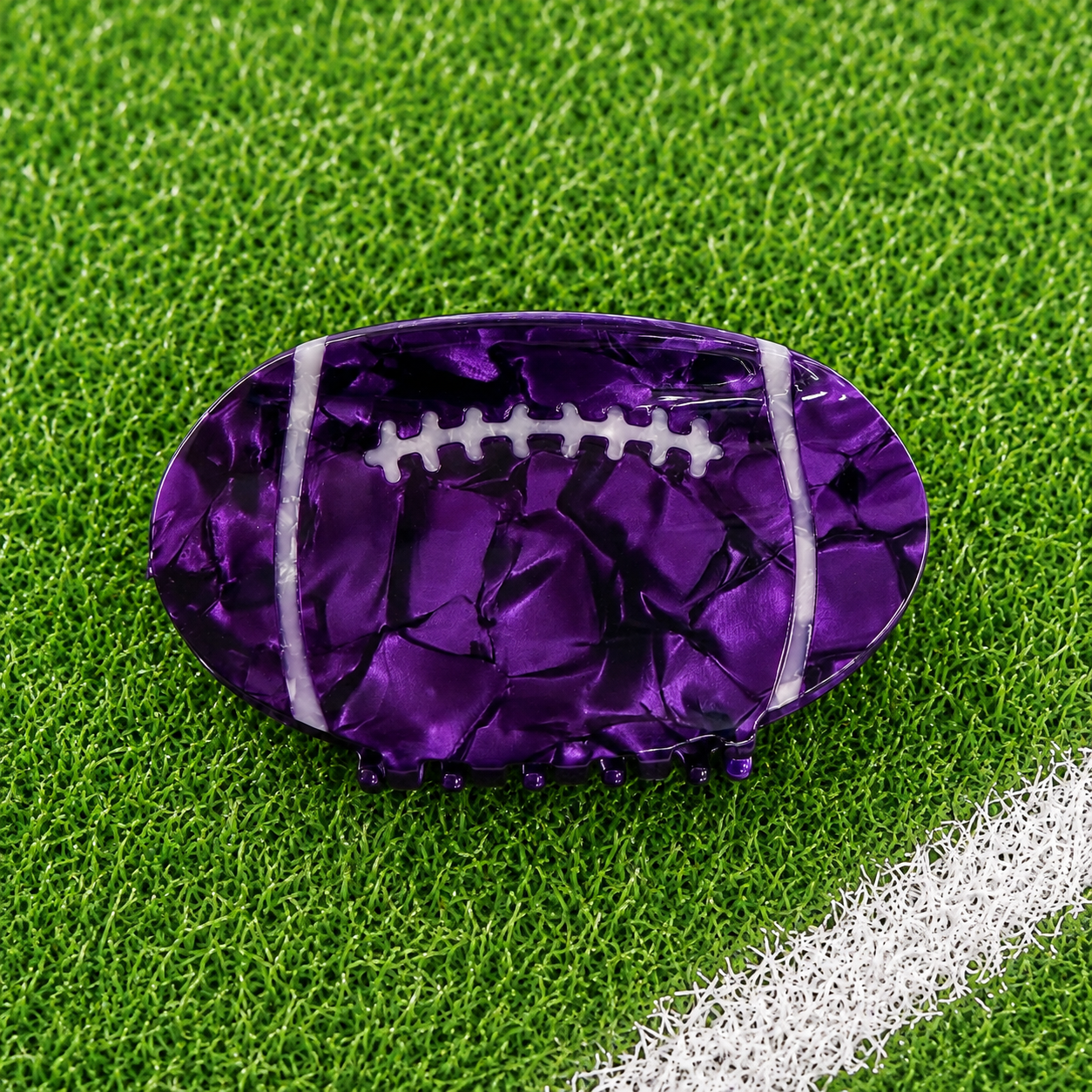 A purple football-shaped accessory with a glossy, cracked metallic texture and white laces, resting on green artificial turf 