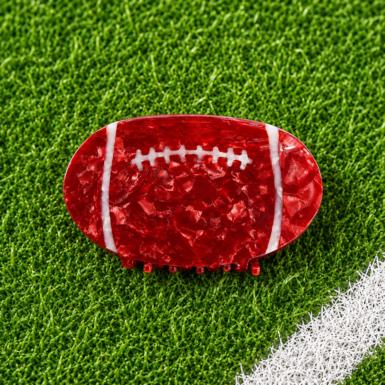 A football-shaped hair claw clip in textured red resin with white laces, displayed on a green artificial turf background.