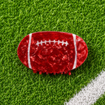 A football-shaped hair claw clip in textured red resin with white laces, displayed on a green artificial turf background.