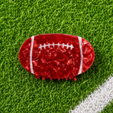 A football-shaped hair claw clip in textured red resin with white laces, displayed on a green artificial turf background.