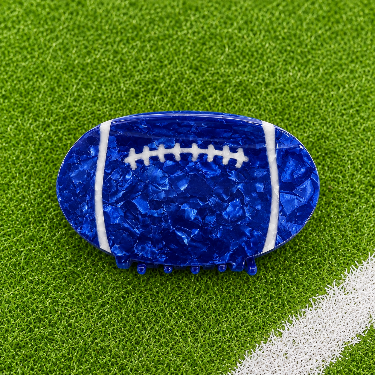A football-shaped hair claw clip in a glossy, textured royal blue resin with white laces, displayed on a green artificial turf background 