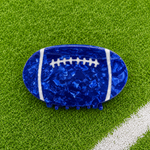 A football-shaped hair claw clip in a glossy, textured royal blue resin with white laces, displayed on a green artificial turf background 