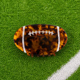 A football-shaped hair claw clip with a brown and black tortoiseshell pattern and white laces, displayed on a green turf background