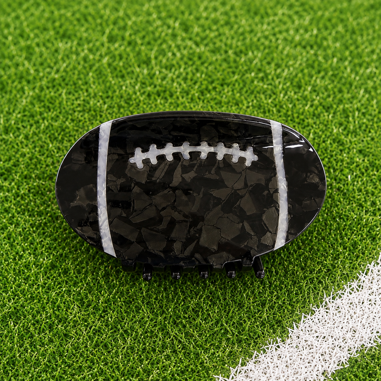 A football-shaped hair claw clip with a glossy black and dark gray marbled pattern, white laces, and white side stripes, displayed on a green turf background