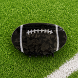 A football-shaped hair claw clip with a glossy black and dark gray marbled pattern, white laces, and white side stripes, displayed on a green turf background