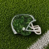 A football helmet-shaped hair claw with a green glittery interior and a clear glittery outline, displayed on a green turf background with a white field line.