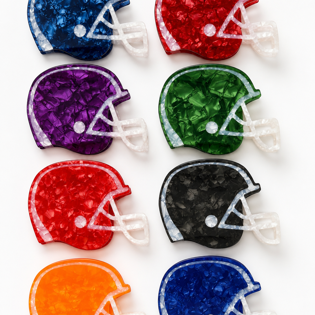 Eight football helmet-shaped hair claws arranged in a 2x4 grid on a white background, each in a different color: navy, red, purple, green, maroon, black, orange, and royal blue. The items have a textured, crackled finish with white faceted details representing laces and faceguard.