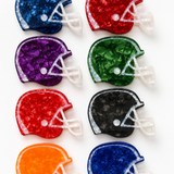 Eight football helmet-shaped hair claws arranged in a 2x4 grid on a white background, each in a different color: navy, red, purple, green, maroon, black, orange, and royal blue. The items have a textured, crackled finish with white faceted details representing laces and faceguard.
