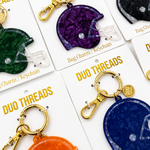 Several football helmet-shaped bag charms and keychains on white cards, each with gold clasp hardware. Visible colors include green, purple, orange, and blue, with the brand name DUO THREADS printed on the cards.