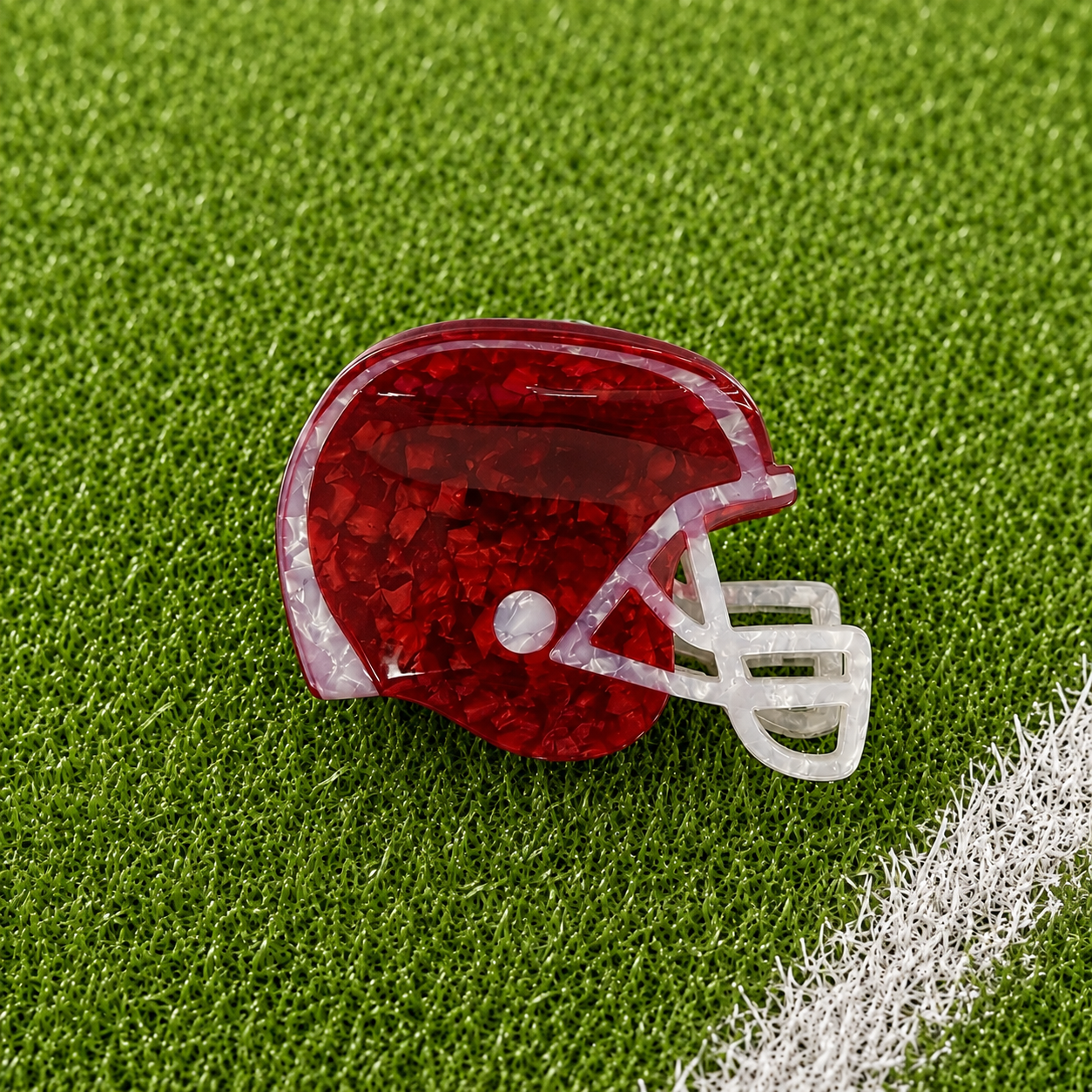 A football helmet-shaped brooch with a maroon glittery shell and a white faceguard, displayed on a green turf background with a white field line.