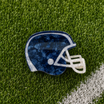 A football helmet-shaped hair claw with a navy blue marbled pattern and a white faceguard, resting on green artificial turf with a white line visible in the corner.