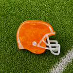 An orange football helmet-shaped hair claw with a translucent white face guard, placed on green artificial turf with a white line visible in the lower right corner.
