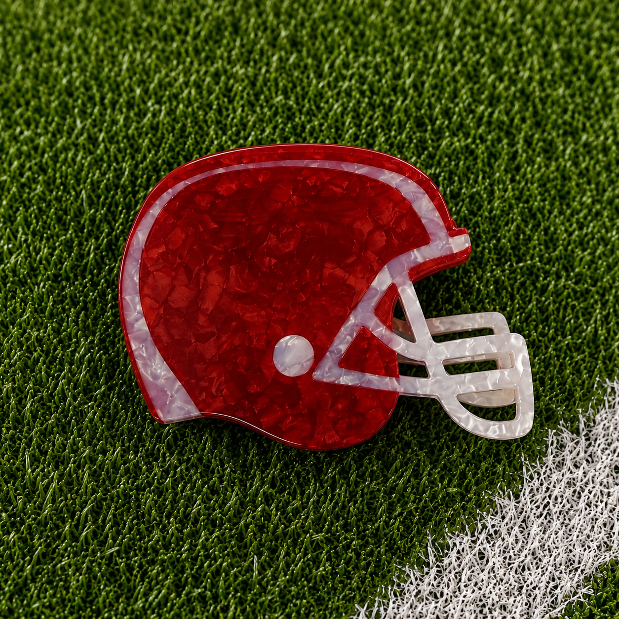 A football helmet-shaped hair claw with a red and white marbled pattern, displayed on a green artificial turf background with a white field line.