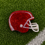 A football helmet-shaped hair claw with a red and white marbled pattern, displayed on a green artificial turf background with a white field line.