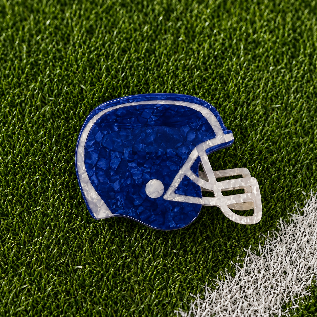 A royal blue and white football helmet-shaped hair claw with a textured, crackled finish, displayed on green artificial turf with a white field line.