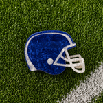 A royal blue and white football helmet-shaped hair claw with a textured, crackled finish, displayed on green artificial turf with a white field line.