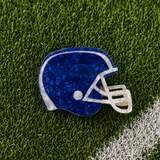 A royal blue and white football helmet-shaped hair claw with a textured, crackled finish, displayed on green artificial turf with a white field line.