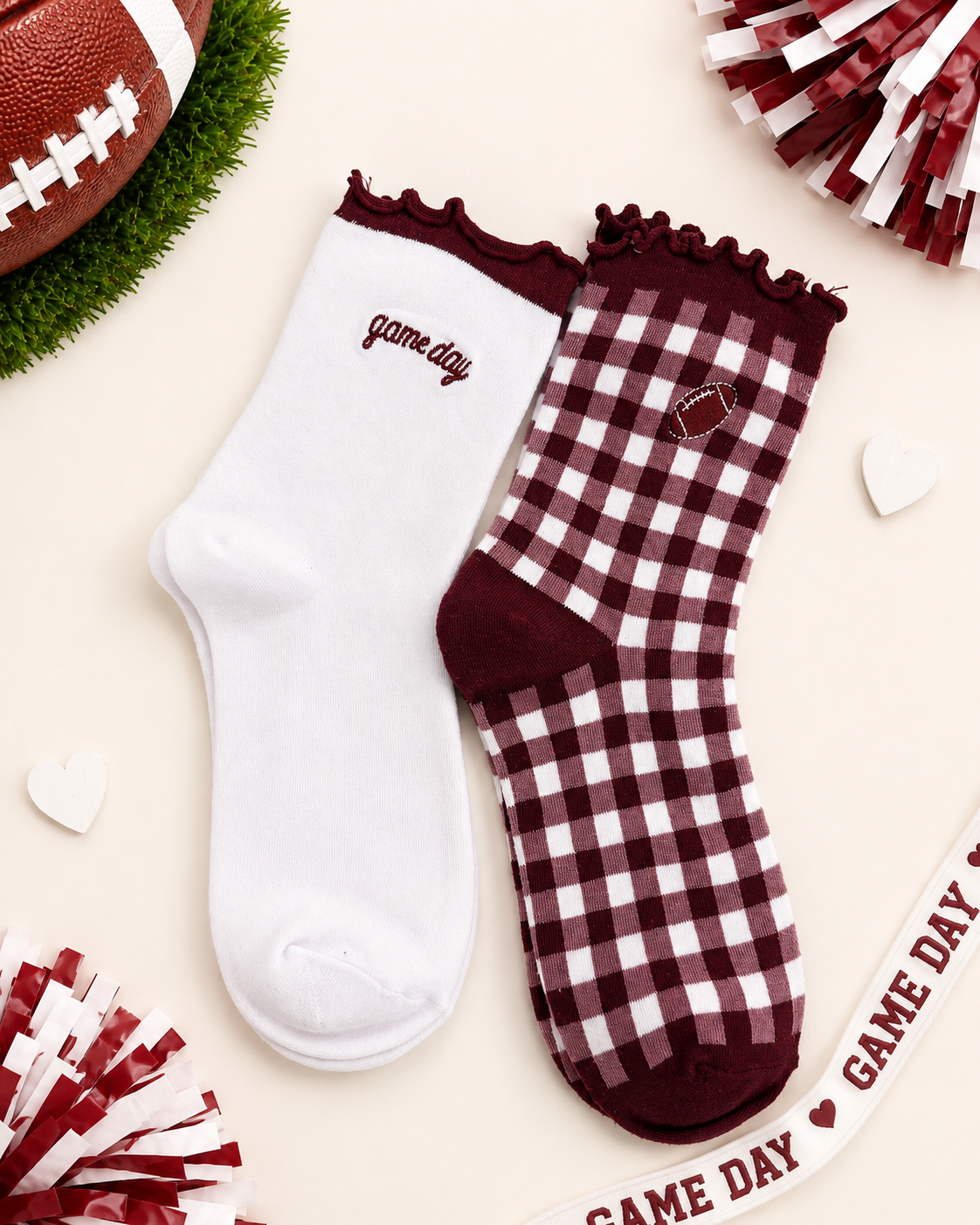 Two ankle socks styled in a flat-lay with football-themed props. One sock is solid white with 'game day' embroidered in burgundy, and the other is a burgundy and white gingham check with a maroon heel, toe, and collar, featuring a small embroidered football patch.