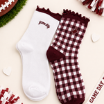 Two ankle socks styled in a flat-lay with football-themed props. One sock is solid white with 'game day' embroidered in burgundy, and the other is a burgundy and white gingham check with a maroon heel, toe, and collar, featuring a small embroidered football patch.