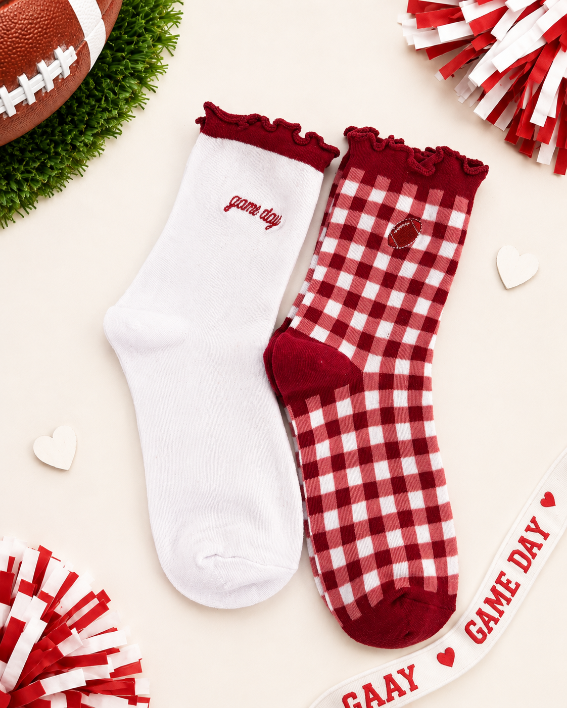 Two decorative ankle socks laid flat on a light cream background with football-themed props. One sock is solid white with scarlet ruffle trim and the word 'game day' embroidered on the side.