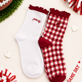 Two decorative ankle socks laid flat on a light cream background with football-themed props. One sock is solid white with scarlet ruffle trim and the word 'game day' embroidered on the side.