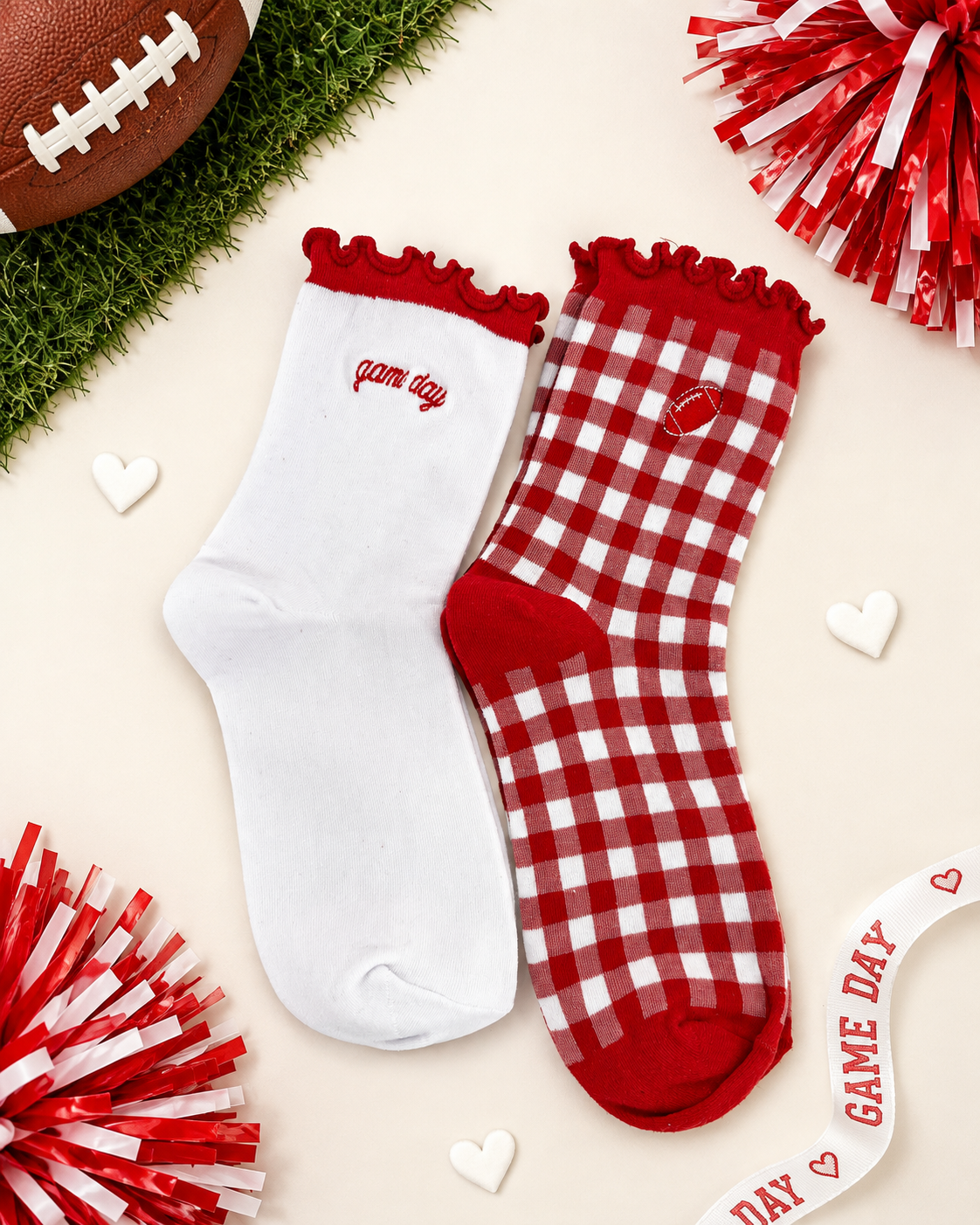 Two red and white socks styled on a light beige background with football and pom-pom props. One sock is solid white with red trim and 'game day' text, and the other is red and white gingham with a small heart patch and red trim.
