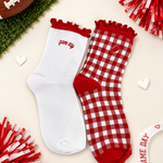 Two red and white socks styled on a light beige background with football and pom-pom props. One sock is solid white with red trim and 'game day' text, and the other is red and white gingham with a small heart patch and red trim.