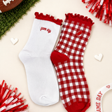 Two red and white socks styled on a light beige background with football and pom-pom props. One sock is solid white with red trim and 'game day' text, and the other is red and white gingham with a small heart patch and red trim.