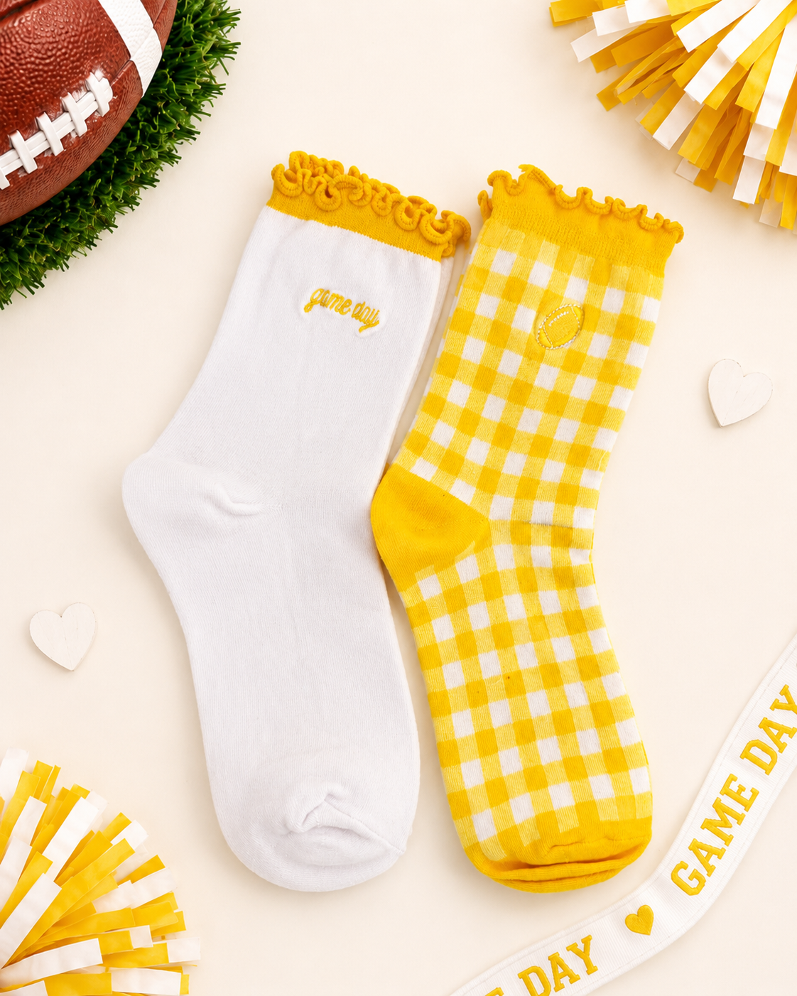 Two socks displayed flat on a light cream background with football-themed props. One sock is white with a yellow ruffled cuff and small yellow 'game day' text; the other is yellow-and-white gingham with a yellow cuff and yellow toe/heel.