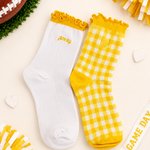 Two socks displayed flat on a light cream background with football-themed props. One sock is white with a yellow ruffled cuff and small yellow 'game day' text; the other is yellow-and-white gingham with a yellow cuff and yellow toe/heel.