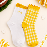 Two socks displayed flat on a light cream background with football-themed props. One sock is white with a yellow ruffled cuff and small yellow 'game day' text; the other is yellow-and-white gingham with a yellow cuff and yellow toe/heel.